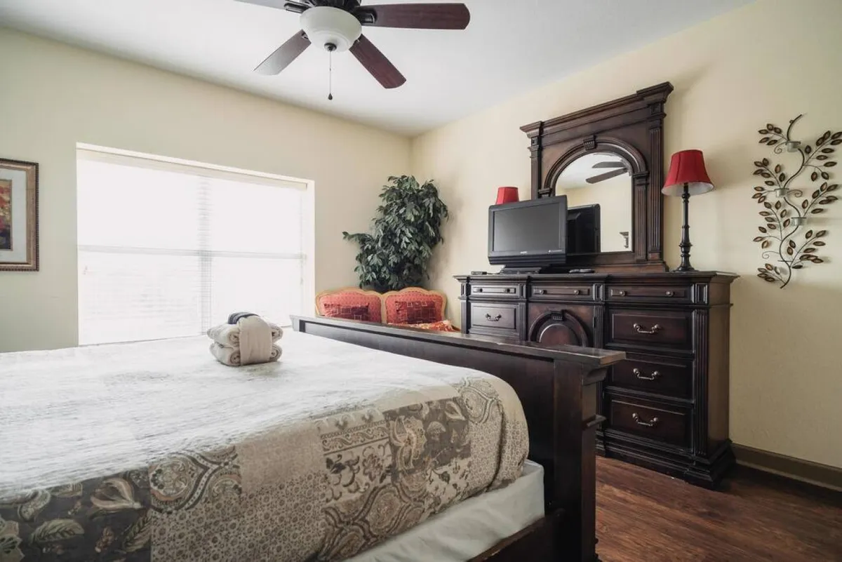 A spacious bedroom featuring a large bed with soft linens, a dresser, an armchair, and a window bringing in natural light. The room is styled with inviting décor and includes a television, blending relaxation and functionality.