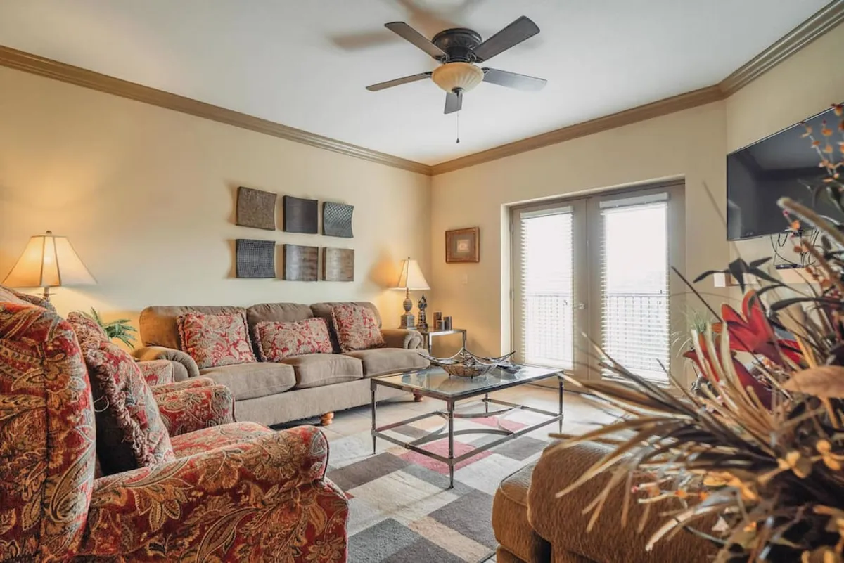 A cozy living room featuring a comfortable sofa with patterned cushions, a decorative armchair, a coffee table, and lamps. Sunlight streams in through the window, and a television sits ready for evening entertainment. The space is styled for relaxation and socializing, offering ample seating for group activities.