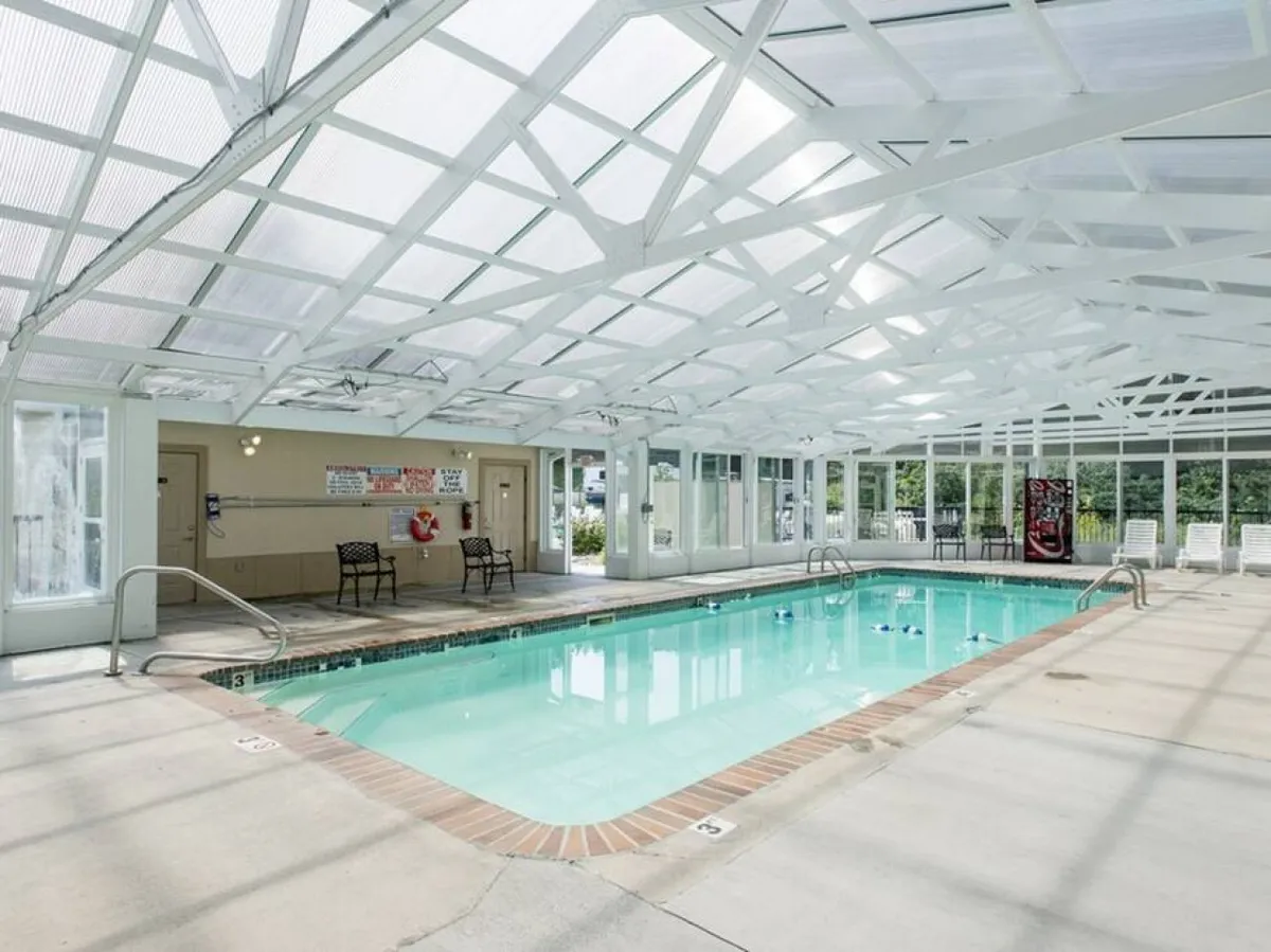 This image shows a bright indoor swimming pool with white ceiling beams and several chairs arranged along the poolside. The space looks inviting and well-maintained, ideal for year-round swimming and social gatherings.