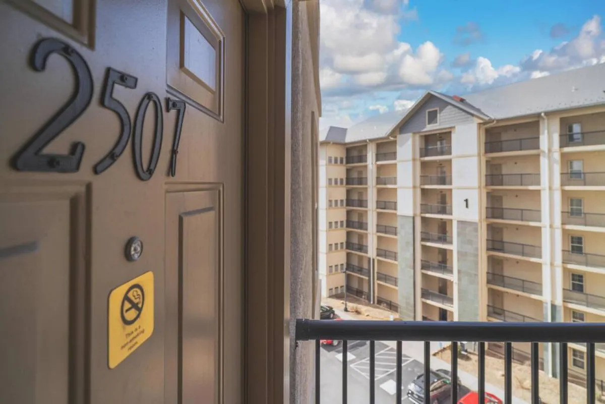 A view from the balcony or entryway, showing the exterior of the building, door number 2507, and hints of outdoor gathering spaces. The image evokes a sense of arrival and the potential for guests to enjoy fresh air and socialize just outside their unit.