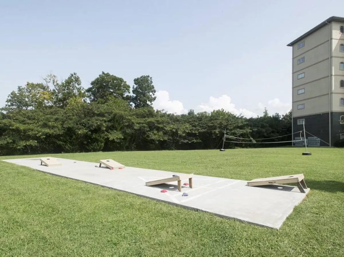 A lively exterior scene with cornhole boards set up on green grass, a building in the background, and a volleyball net visible. The area feels open and communal, ideal for games, picnics, and group activities.