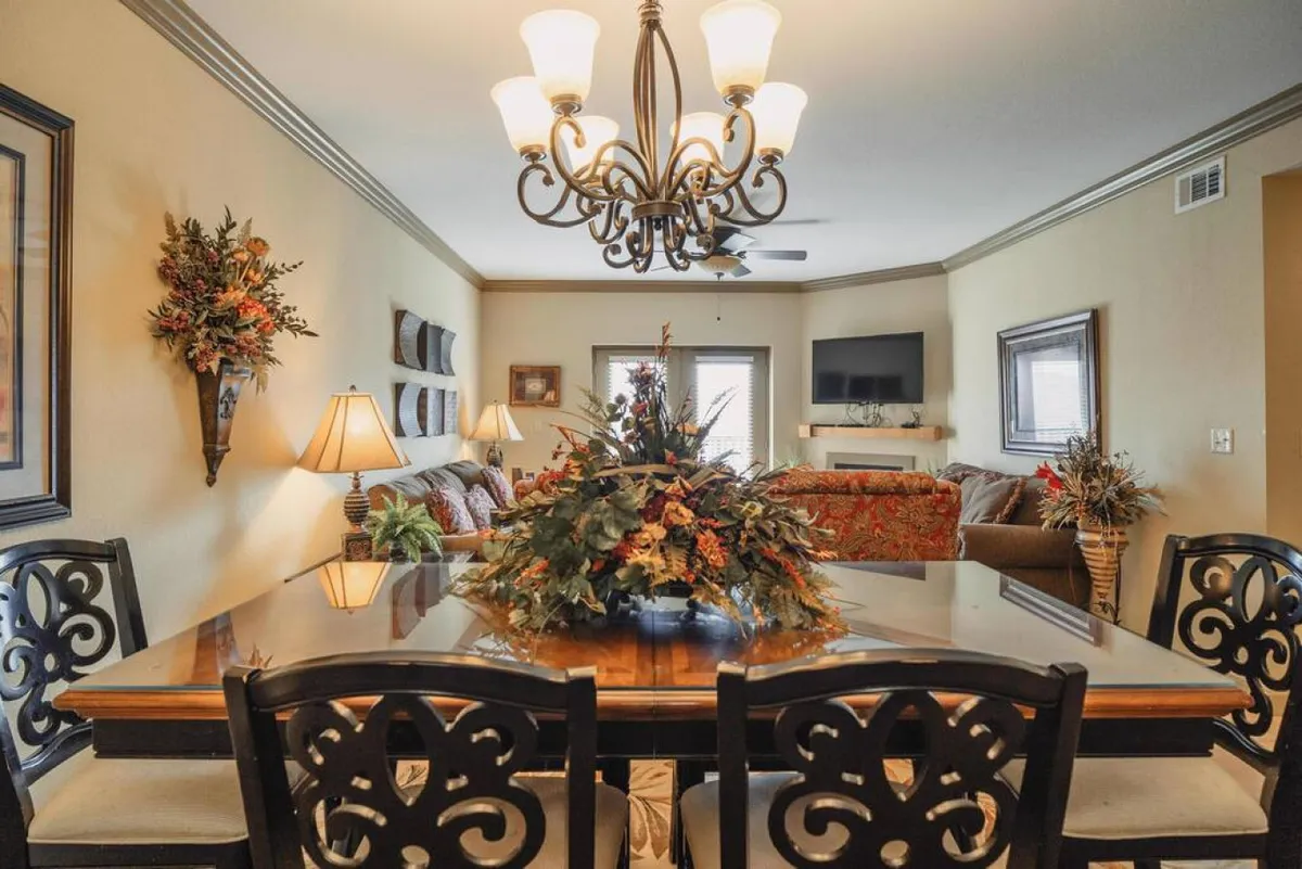A spacious open-concept setting with a dining table adorned with a floral centerpiece, an elegant chandelier overhead, tasteful wall decor, and a view extending into the living room seating area. The room is bathed in natural light, highlighting both the dining and social spaces.