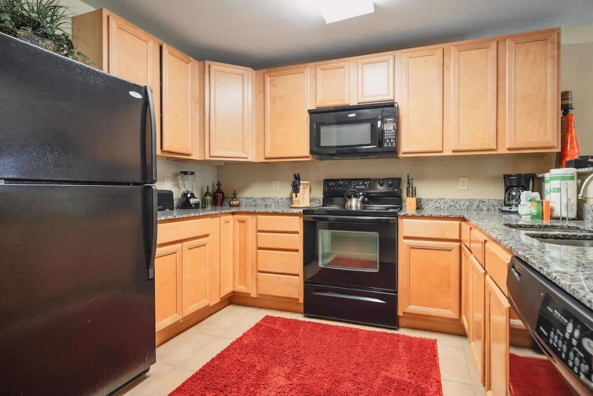 A close-up view of the kitchen area, featuring a black refrigerator, granite countertops, a cooking range, microwave, and a red rug. The scene feels cozy yet highly functional, emphasizing the availability of all essential appliances for meal prep.