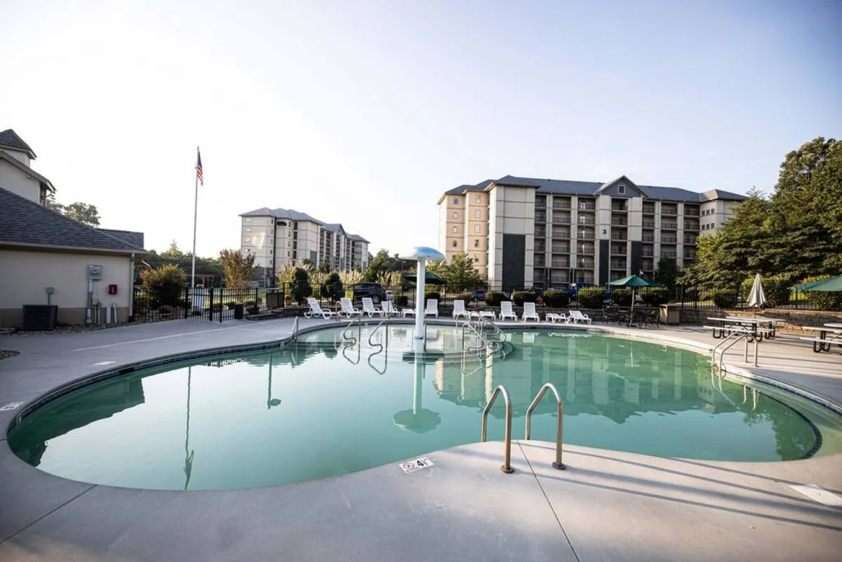 An inviting outdoor swimming pool surrounded by lounge chairs and green umbrellas, set against a backdrop of lush trees and a flag pole, evoking a lively yet relaxed atmosphere for family fun and social gatherings.