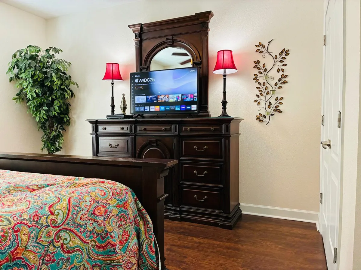 The image showcases a cozy and inviting bedroom with a colorful bedspread, a decorative dresser, a TV and mirror setup, a potted plant, and tasteful wall decor. The scene suggests a comfortable space for relaxation, movie nights, or quiet mornings.