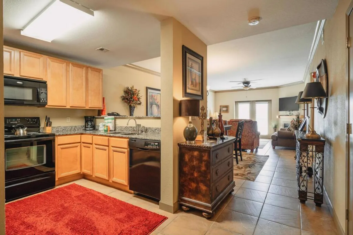 This perspective captures the kitchen cabinets, red rug, adjacent dining area, and a glimpse of the cozy living room. Decorative items add warmth, while the open flow between the kitchen and dining/living spaces suggests easy connection for group meals.
