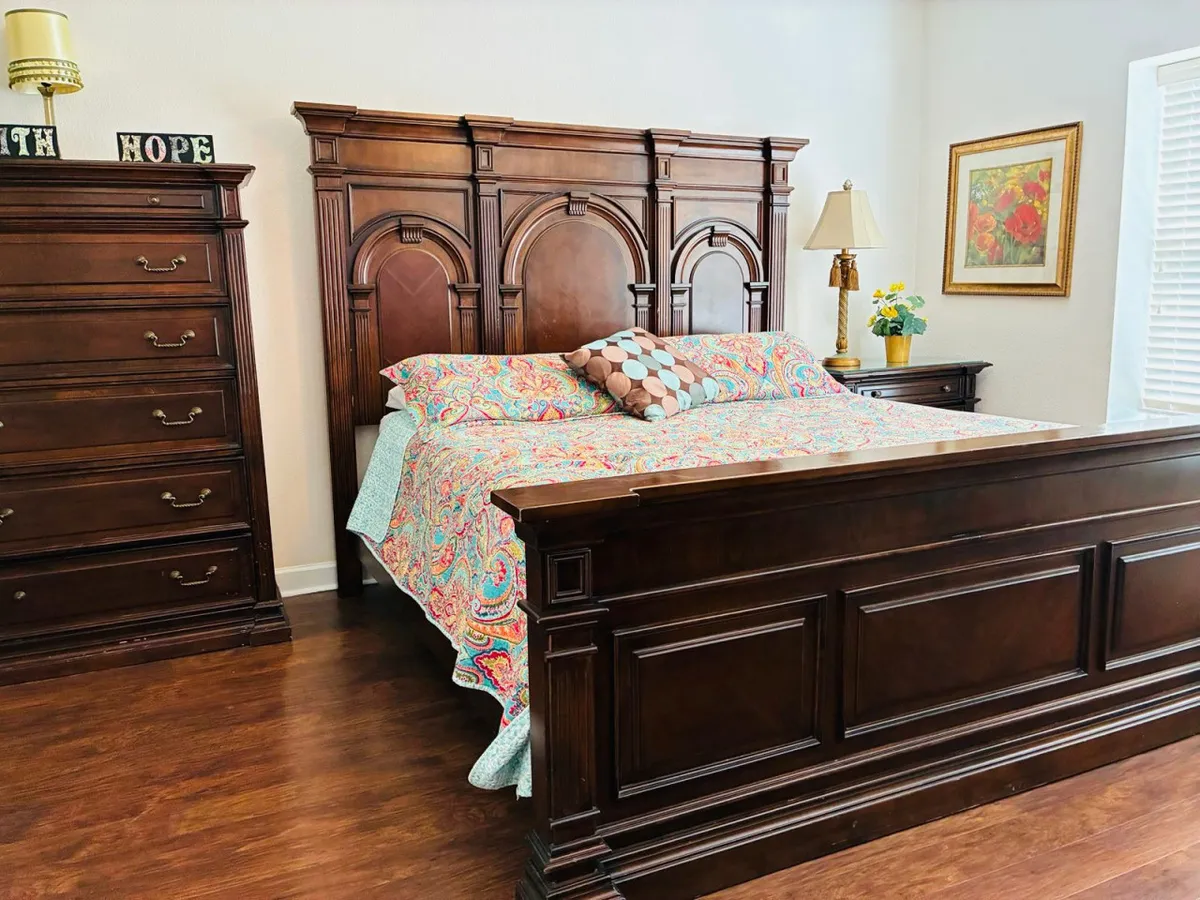A cozy bedroom with an ornate headboard, colorful bedspread, decorative pillows, a nightstand with lamp, and floral artwork on the wall. The inviting design suggests a comfortable and private retreat, perfect for unwinding.