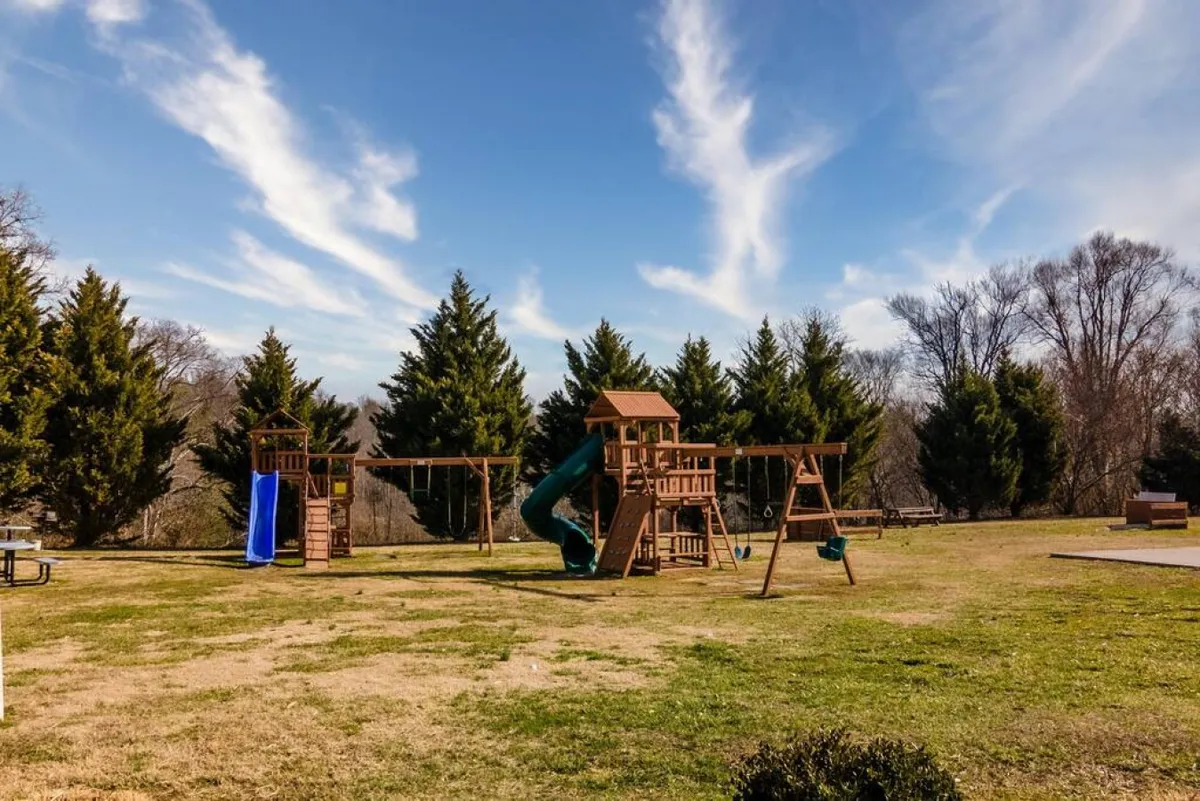 This image shows a colorful playground structure with swings and a slide, set in a grassy area fringed with trees. Children and families can be seen enjoying the space, emphasizing a playful and inviting environment.